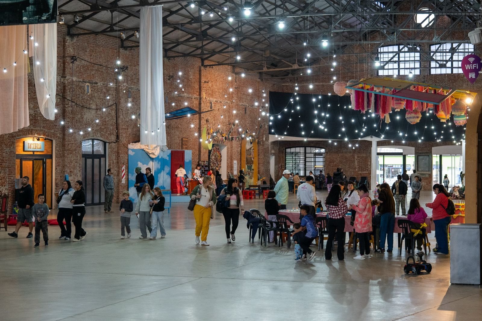 Familias recorren un galpón de ladrillo con guirnaldas de luces y mesas de actividades